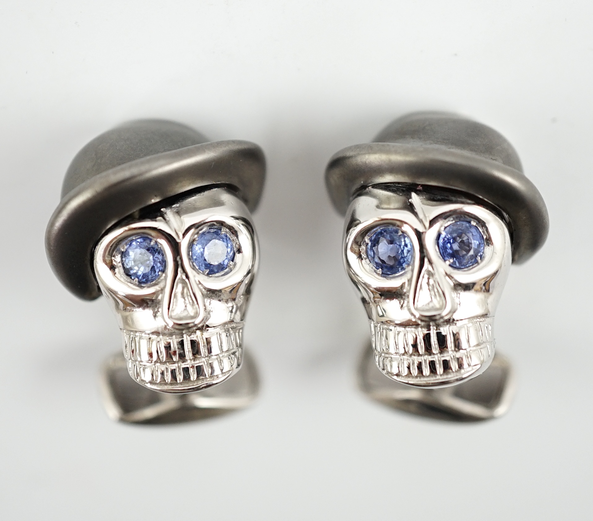 A modern pair of Longmire 18ct white gold, blackened rhodium and sapphire set novelty cufflinks, each modelled as a skull wearing a bowler hat